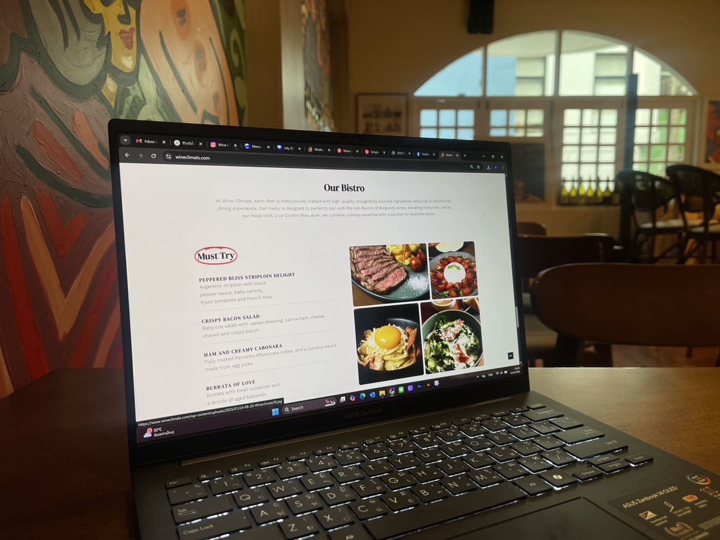 หน้าจอแล็ปท็อปแสดงเว็บไซต์ Wine Climats หน้า Our Bistro พร้อมเมนูอาหาร Must Try รวมถึงภาพอาหารสวยงาม ตั้งอยู่ในร้านอาหารบรรยากาศอบอุ่น เป็นตัวอย่างผลงานรับทำเว็บไซต์ร้านอาหารและไวน์บาร์